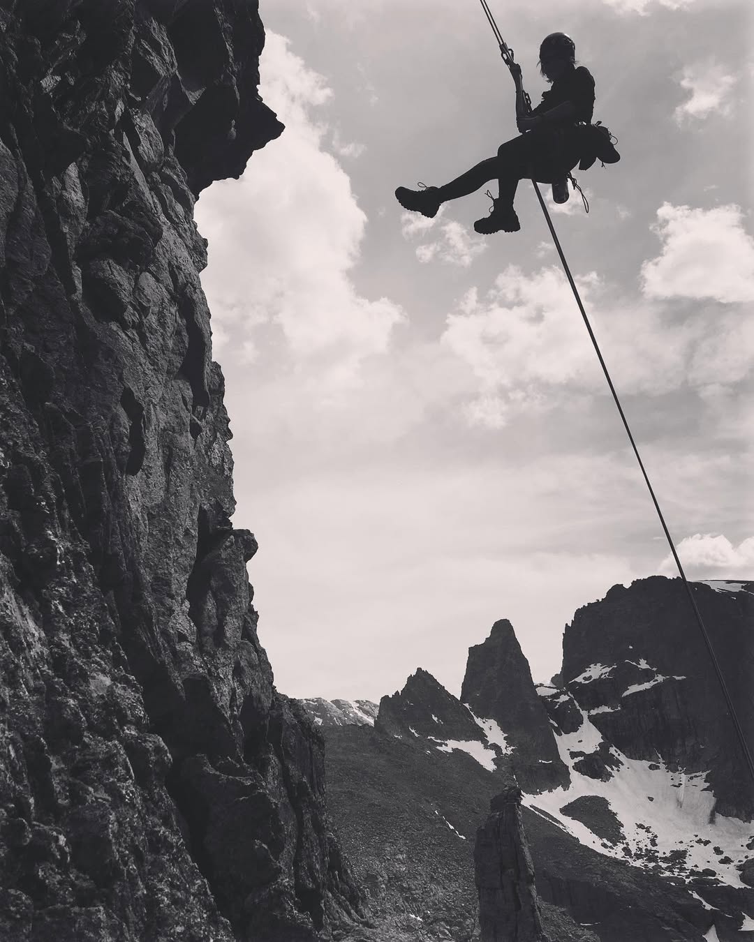 Black and white photo shows a female climber rappelling off of Wham, a high alpine climb in RMNP. She is silhouetted against the sky , and the rope trails away towards the ground. In the background, the top of Zowie tower is visible, as is the Sharkstooth, far-away.